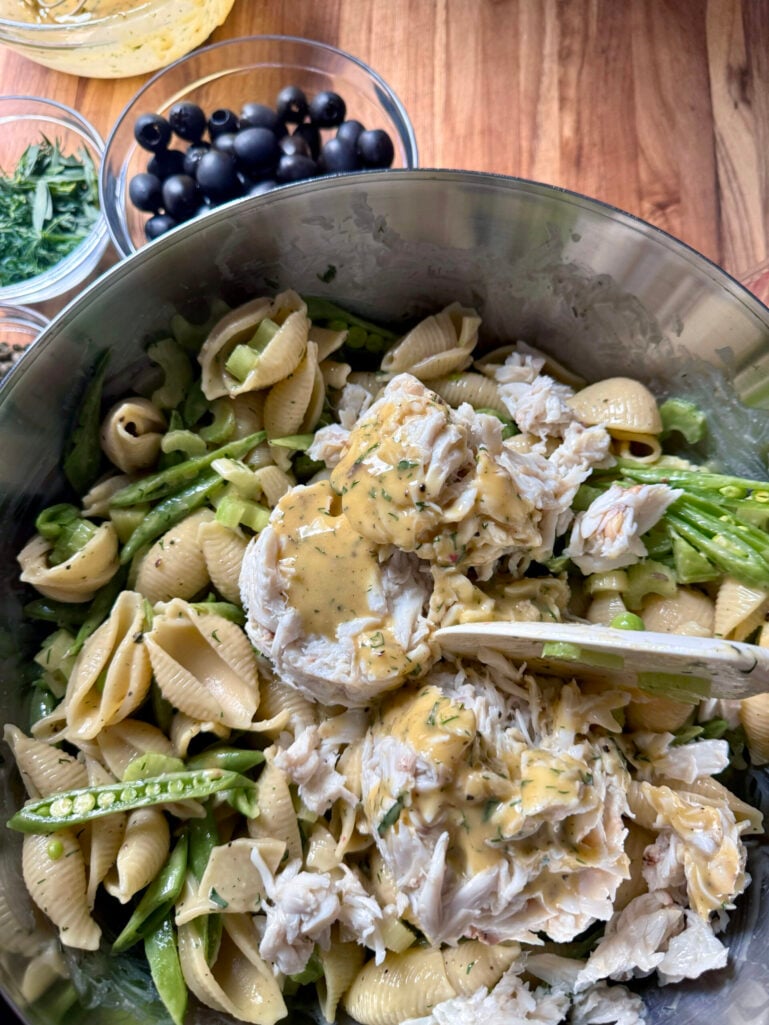 Lump crab meat being folded into the pasta, snap pea, and celery mixture in a large stainless bowl