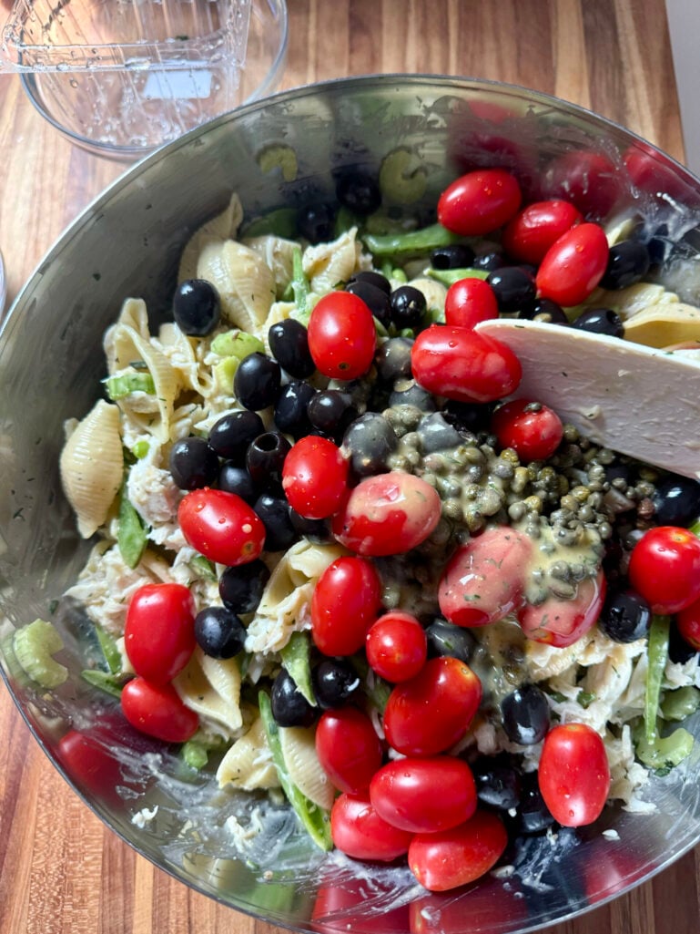 Grape tomatoes, black olives, and capers added on top of the crab and pasta salad in a large stainless mixing bowl