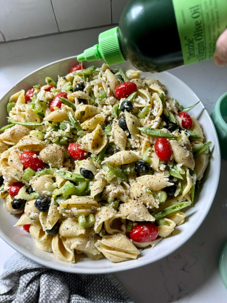 Finished seafood pasta salad in a large white serving bowl being drizzled with Grazia extra virgin olive oil, with a small bowl of flaky salt and fresh herbs alongside