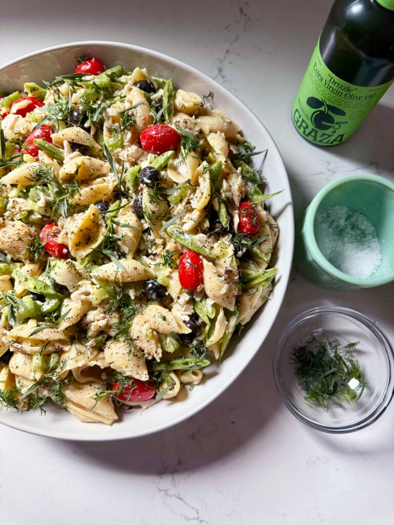 Overhead view of the finished seafood pasta salad in a large white bowl, garnished with fresh dill and tarragon, with Grazia olive oil and a cup of flaky salt alongside on a marble counter