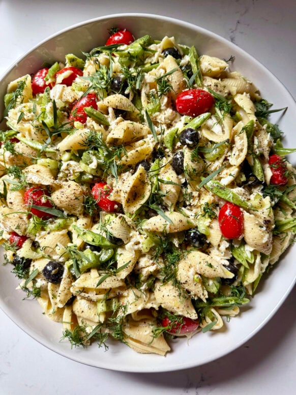 Close-up overhead of the finished seafood pasta salad garnished with fresh dill and tarragon, showing pasta shells, grape tomatoes, black olives, snap peas, celery, and lump crab