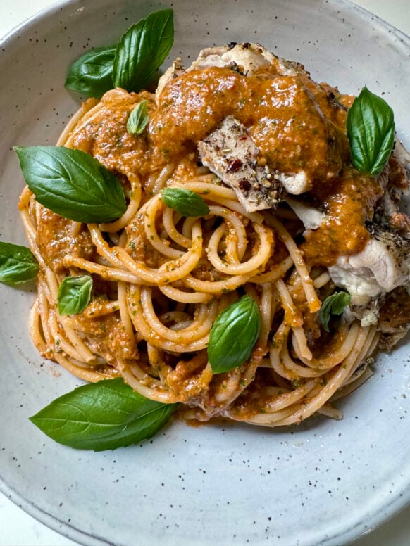 Overhead close-up of spaghetti tossed in pesto pantesco topped with sliced baked chicken and fresh basil leaves in a speckled ceramic bowl