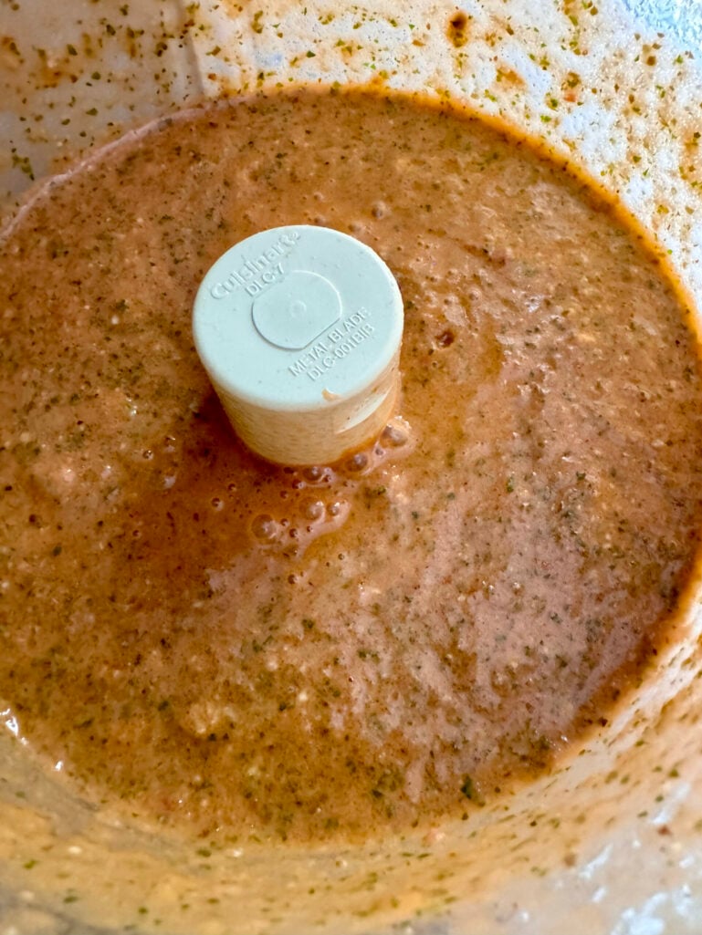 Close-up of the finished pesto pantesco in the food processor bowl, showing a smooth, rusty-orange sauce with flecks of herbs