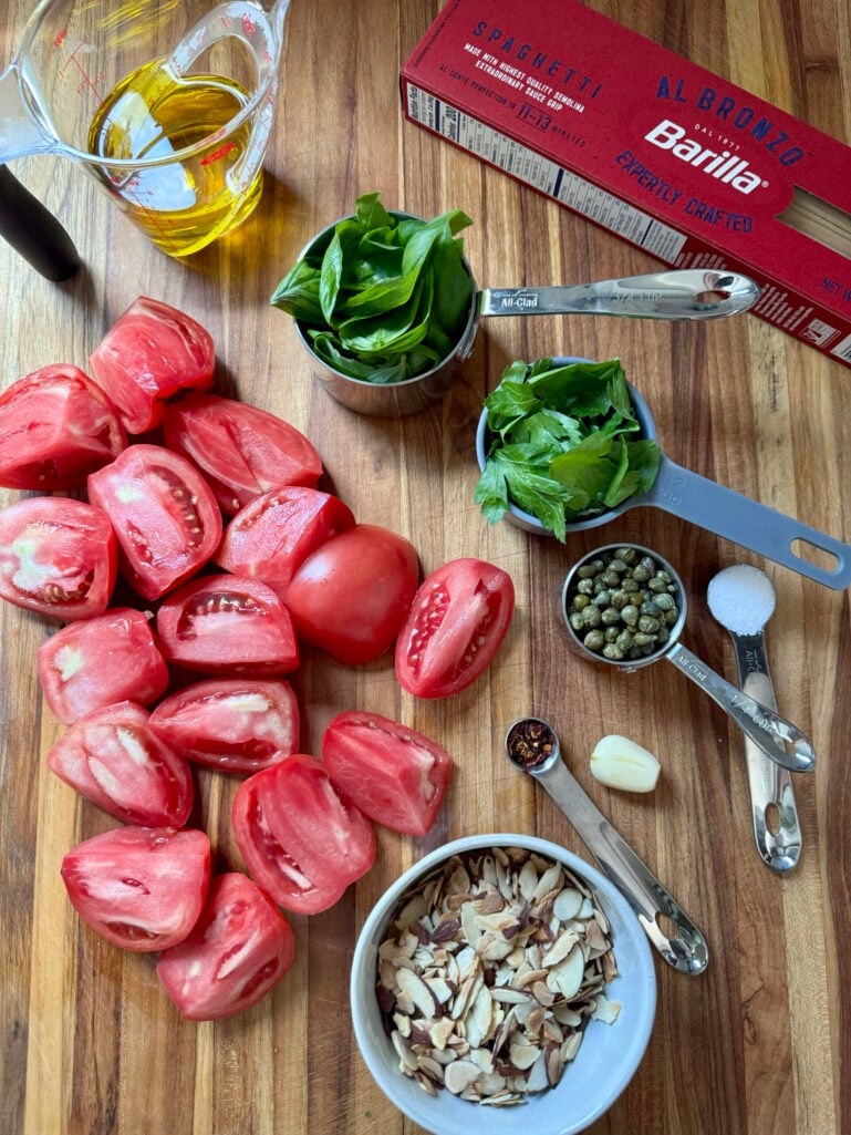 Overhead view of ingredients on a wooden cutting board: quartered fresh tomatoes, fresh basil and parsley, toasted slivered almonds, capers, garlic clove, red pepper flakes, kosher salt, olive oil, and a box of Barilla al Bronzo spaghetti