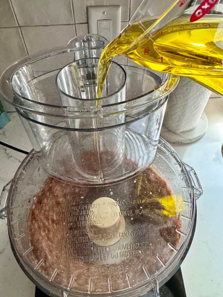Olive oil being poured from a measuring cup through the feed tube of a Cuisinart food processor while blending the pesto pantesco