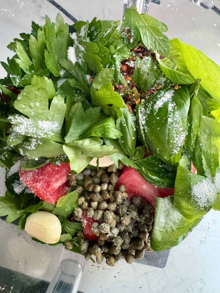 Overhead view inside the food processor bowl with quartered tomatoes, fresh basil, parsley, capers, garlic, salt, and red pepper flakes before blending