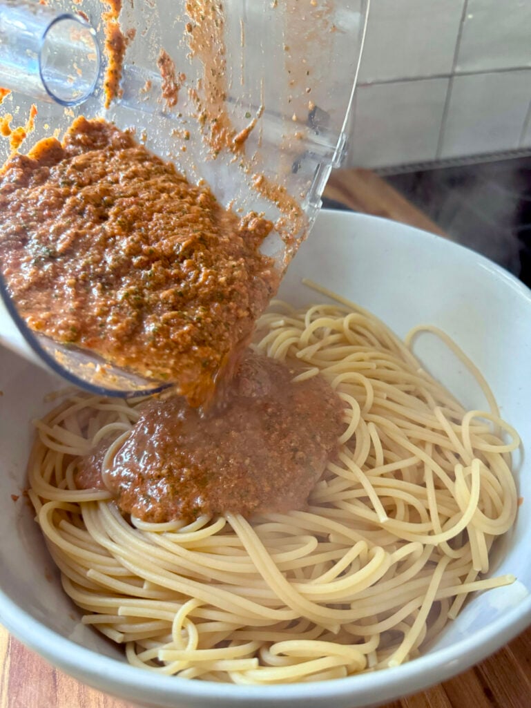 Pesto pantesco being poured from the food processor bowl over cooked spaghetti in a white serving bow