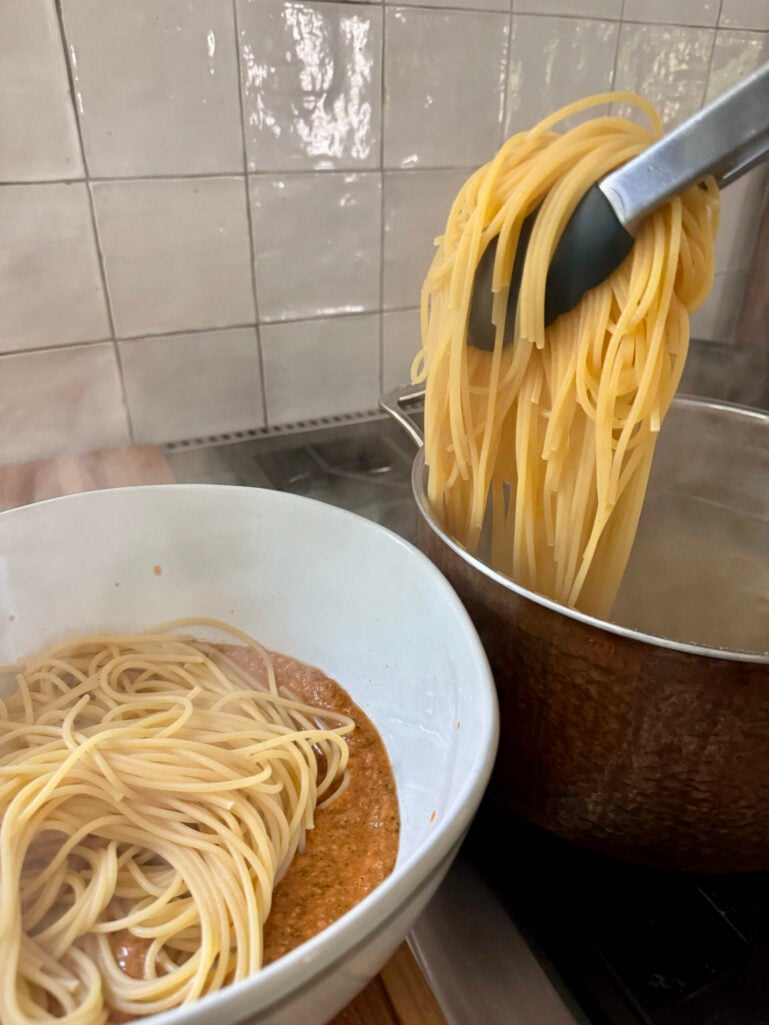 Tongs transferring cooked spaghetti from the pot into a large white serving bowl with pesto pantesco