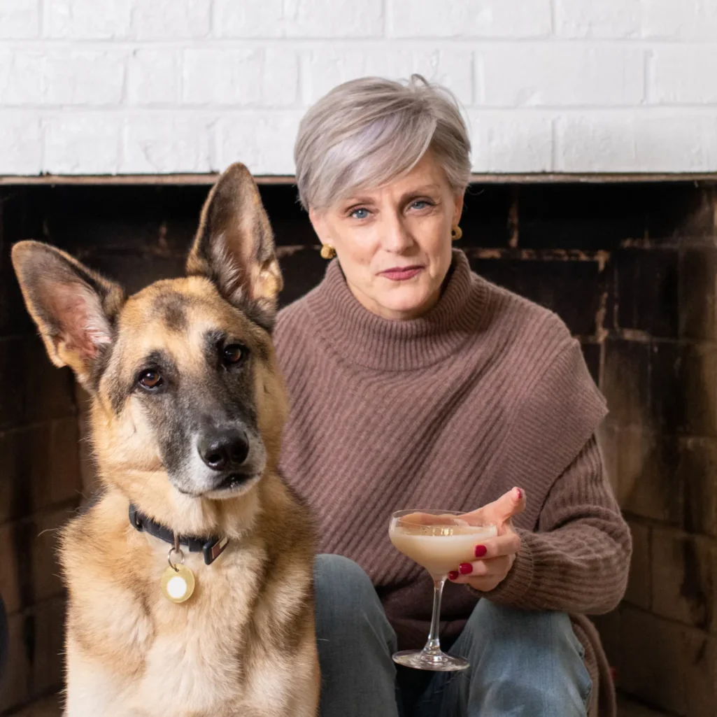 Beth in brown turtleneck holding cocktail by fireplace with Oscar the German Shepherd with distinguished grey muzzle - Fridays with Oscar