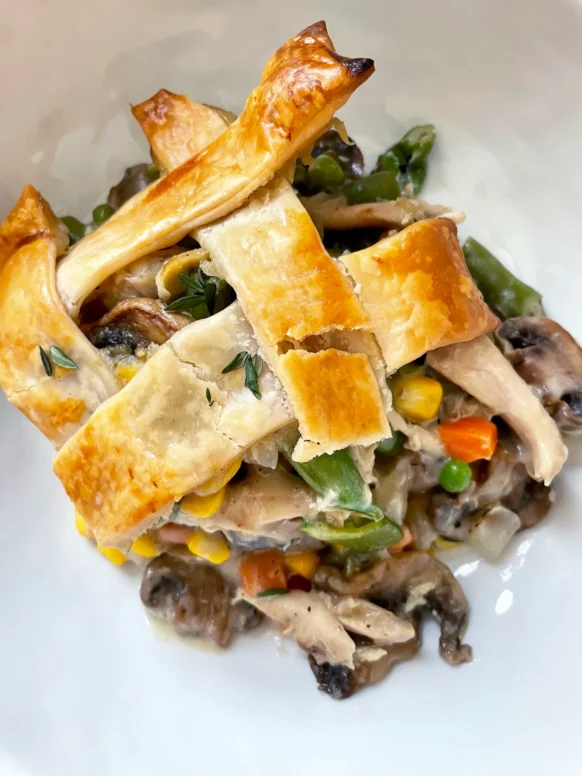 Close-up of a serving of spring chicken pot pie in a white bowl showing golden lattice crust strips over creamy filling with mushrooms, corn, peas, carrots, and shredded chicken, garnished with fresh thyme