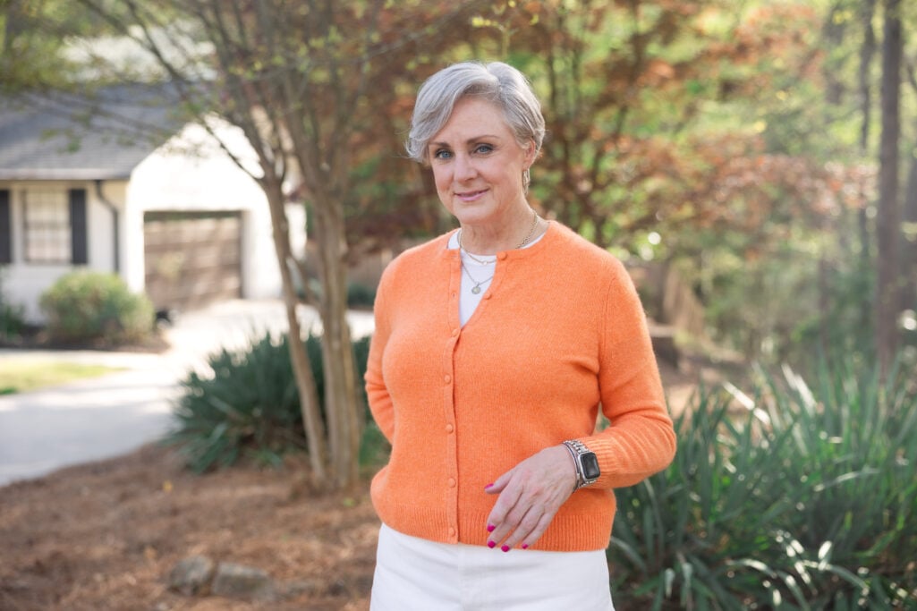 Woman over 50 wearing bright orange cardigan with white jeans.