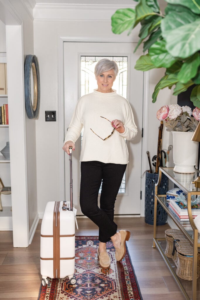 Beth Djalali in ivory sweater, black pants, and loafers holding white suitcase—chic fall travel outfit for women over 50.