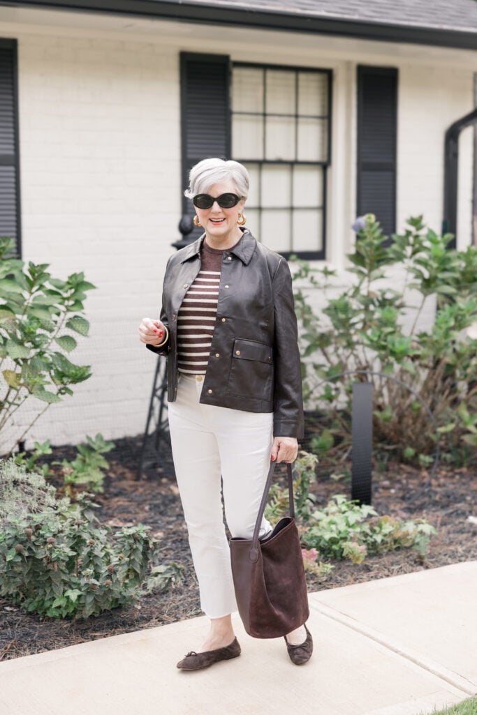 Beth Djalali walking confidently on Tybee Island in a brown faux leather jacket, striped sweater, ivory jeans, and brown suede flats, holding a matching hobo bag in front of a white-brick cottage.