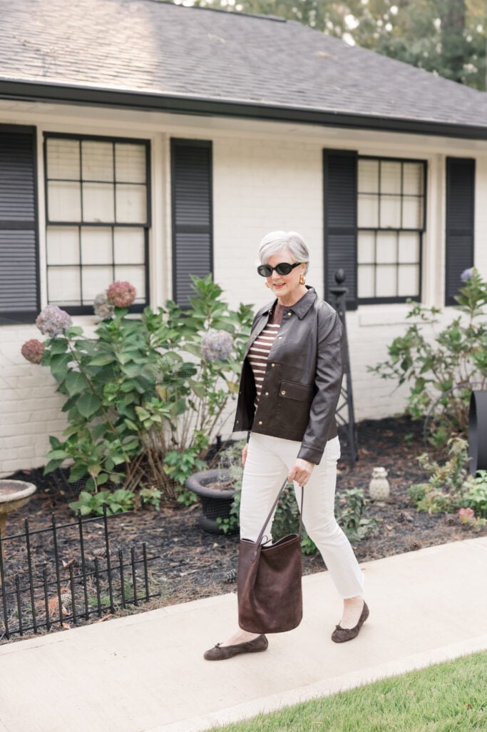 Beth Djalali walking confidently on Tybee Island in a brown faux leather jacket, striped sweater, ivory jeans, and brown suede flats, holding a matching hobo bag in front of a white-brick cottage.