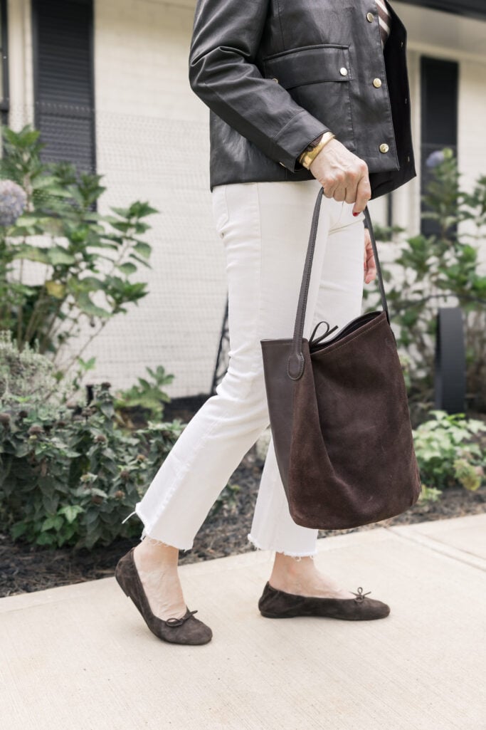 Beth Djalali walking confidently on Tybee Island in a brown faux leather jacket, striped sweater, ivory jeans, and brown suede flats, holding a matching hobo bag in front of a white-brick cottage.