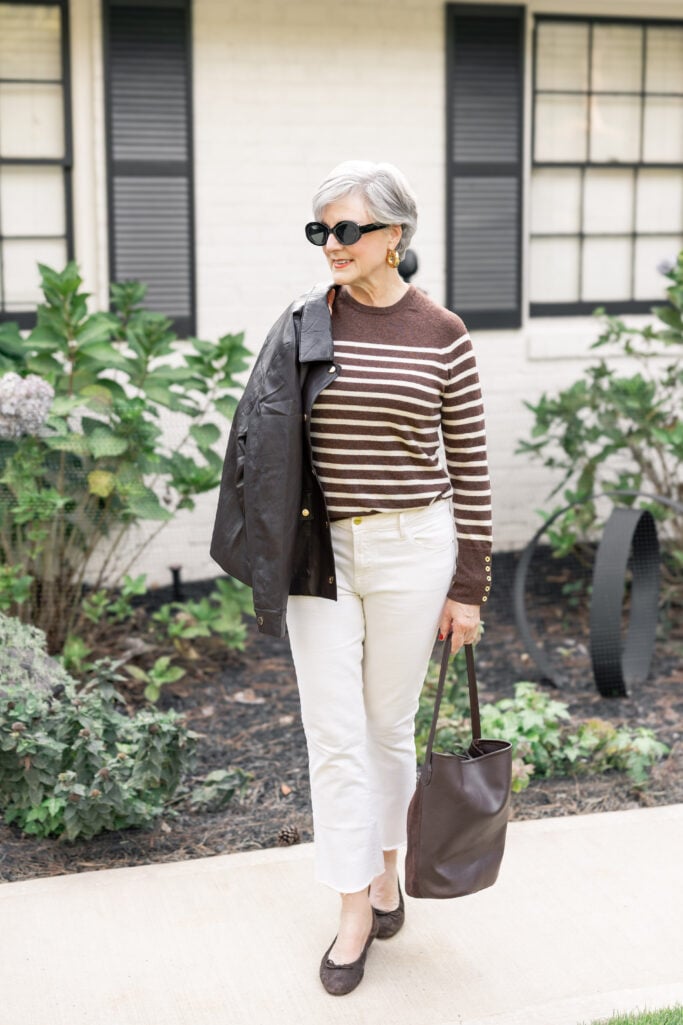 Beth Djalali walking confidently on Tybee Island in a brown faux leather jacket, striped sweater, ivory jeans, and brown suede flats, holding a matching hobo bag in front of a white-brick cottage.