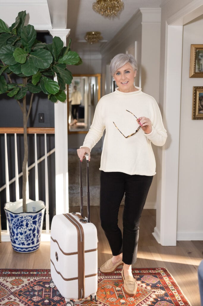 Beth Djalali wears a cream sweater with black ankle pants and loafers, holding a white suitcase — casual travel style over 60.
