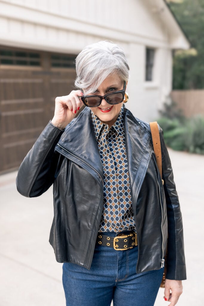 Woman Looking over her sunglasses wearing dark wash jeans and a leather jacket.