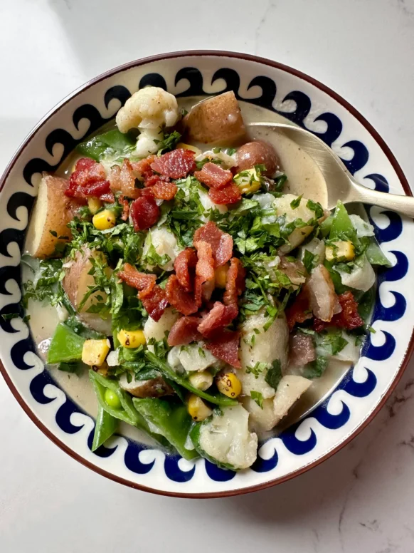 Bowl of dairy-free seafood chowder with cod, cauliflower, red potatoes, snow peas, corn, spinach, crispy bacon, and fresh parsley in decorative blue and white bowl