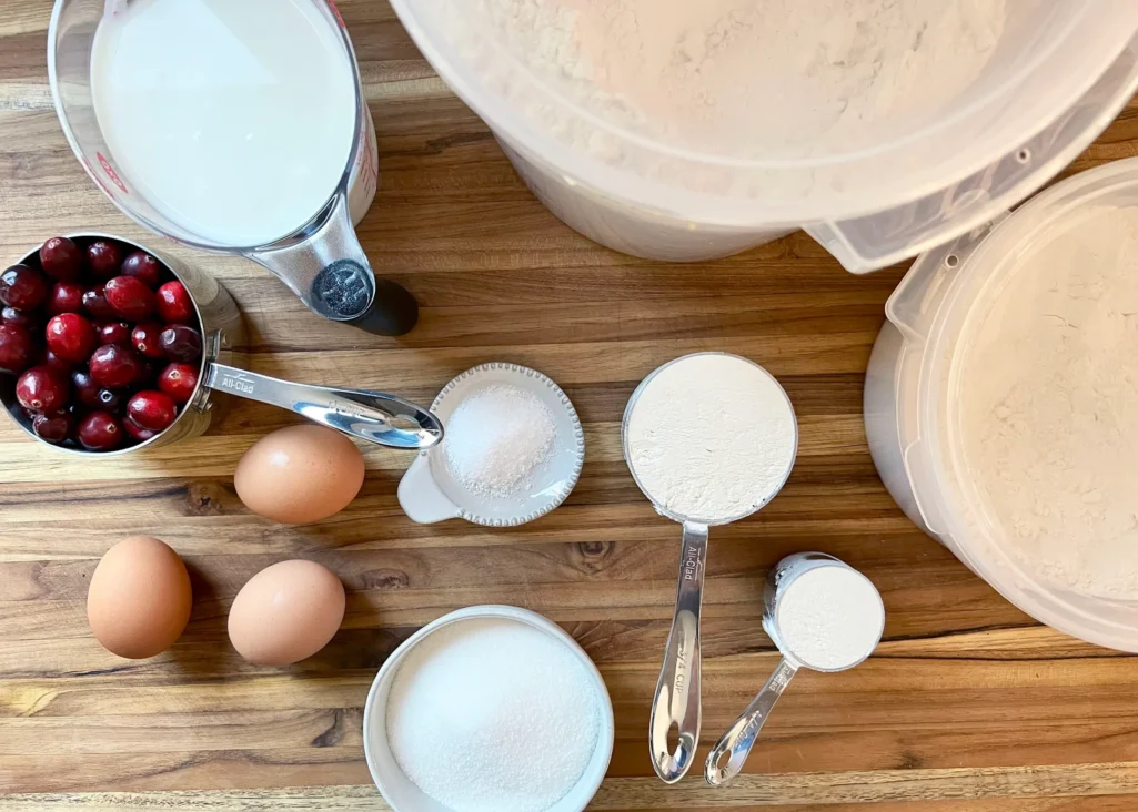 Clafoutis ingredients on a wooden cutting board: milk, eggs, fresh cranberries, sugar, salt, and flour