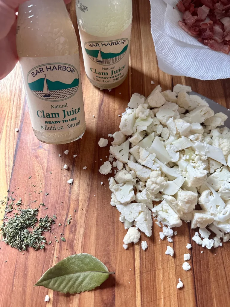 Ingredients for dairy-free seafood chowder including Bar Harbor clam juice bottles, chopped cauliflower, fresh thyme, bay leaf, and chopped bacon on wooden cutting board
