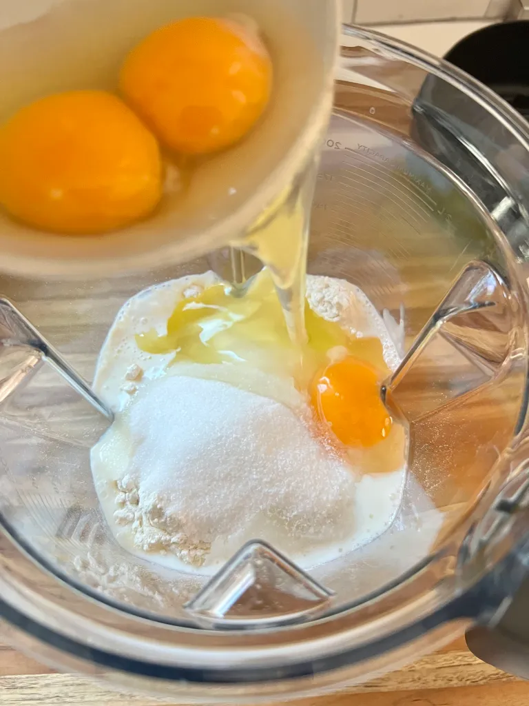 Pouring eggs into a blender with milk, sugar, and flour for clafoutis batter