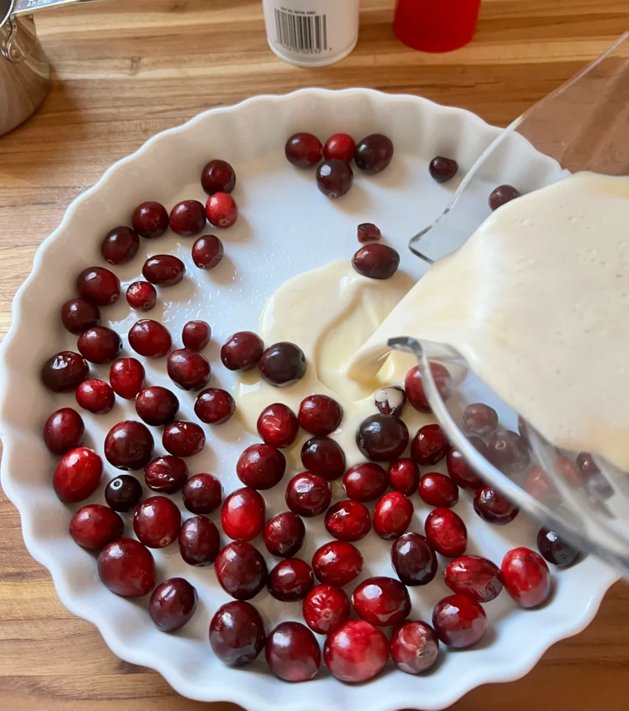 Pouring clafoutis batter from blender over fresh cranberries in a white baking dish