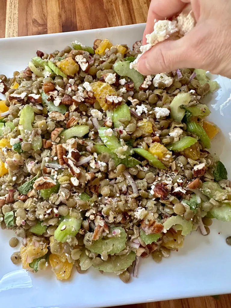 Hand sprinkling feta cheese over plated lentil salad with oranges and pecans - serving healthy lentil salad