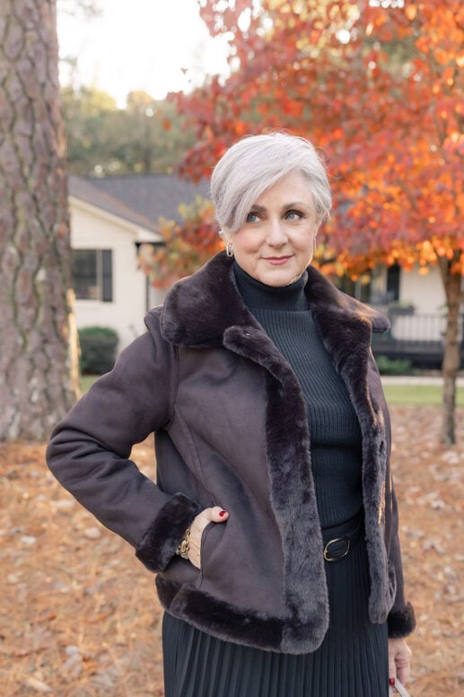 Woman with short silver hair wearing a dark shearling-trimmed jacket over a black ribbed turtleneck and pleated skirt, standing outdoors among fall foliage with orange and red leaves.