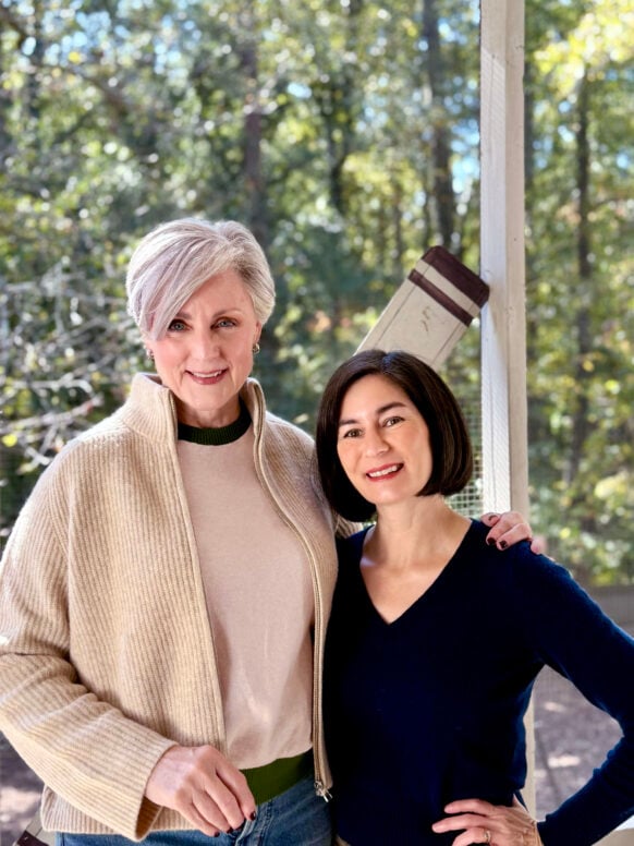 Beth and Kelly smiling together on the porch, dressed in cozy fall sweaters — a beige zip cardigan and a navy V-neck — for a relaxed, sweater-weather entertaining moment.