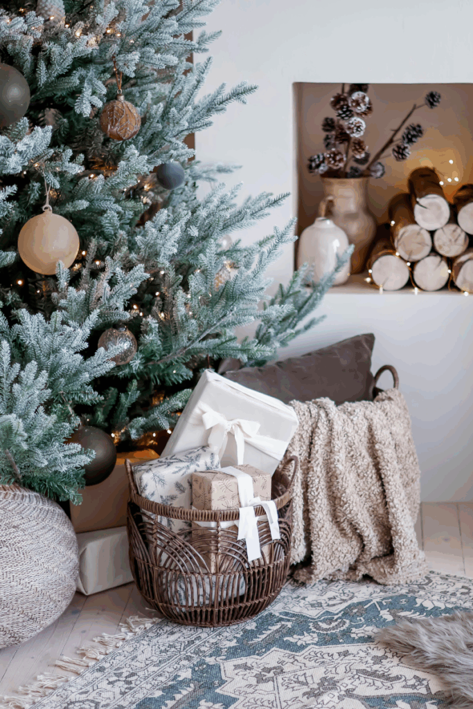 Cozy holiday living room scene with a flocked Christmas tree decorated in neutral ornaments, a wicker basket filled with wrapped gifts tied with white ribbons, a chunky knit throw draped over a basket, stacked firewood wrapped in twinkle lights, and winter greenery arranged in ceramic vases on a built-in shelf.