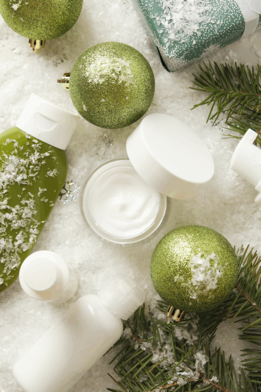 Winter skincare flatlay featuring jars and bottles of cream, lotion, and serum arranged on faux snow with green glitter ornaments, evergreen sprigs, and gift wrap—festive holiday styling for a winter skincare theme.