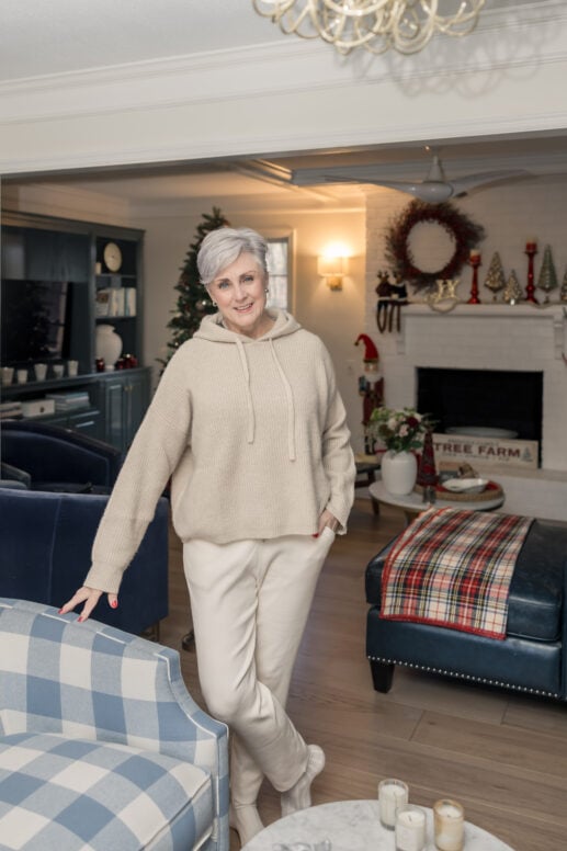 Beth wearing cream ribbed hooded sweater with matching cream trousers and neutral boots, standing in holiday-decorated living room with blue and white buffalo check armchair, navy leather sofa with plaid throw, white fireplace mantel styled with red berry wreath, red candlesticks, decorative Christmas trees, nutcracker, and Tree Farm sign, Christmas tree visible in background near dark blue built-in cabinets
