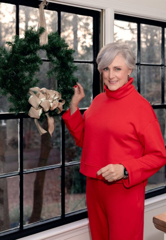 Beth wearing red funnel-neck Spanx top and matching red pants, standing by black-framed windows with evergreen wreath with gold-edged burlap bow, Apple Watch visible