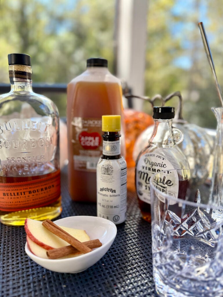 Ingredients for an Apple Cider Old Fashioned cocktail including Bulleit bourbon, apple cider, Angostura bitters, maple syrup, and apple slices arranged on an outdoor table.