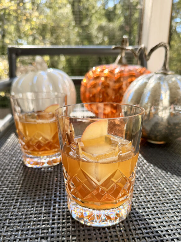 Apple Cider Old Fashioned cocktails garnished with apple slices and cinnamon sticks in crystal glasses, set against a backdrop of metallic pumpkins.