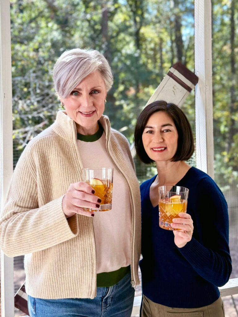 Beth and Kelly cheers with Apple Cider Old Fashioned cocktails while wearing cozy fall sweaters on the screened porch.