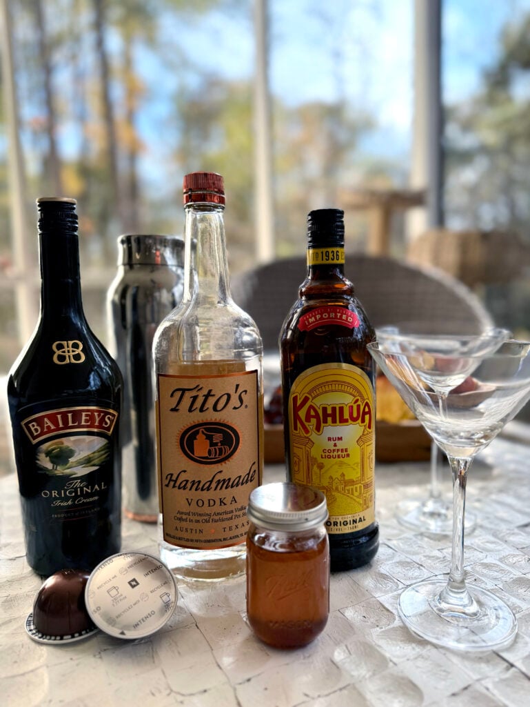 Bar setup with Baileys Irish Cream, Tito's Handmade Vodka, Kahlúa coffee liqueur, Nespresso pods, a small jar of homemade cinnamon syrup, and a martini glass on a marble counter with natural window light