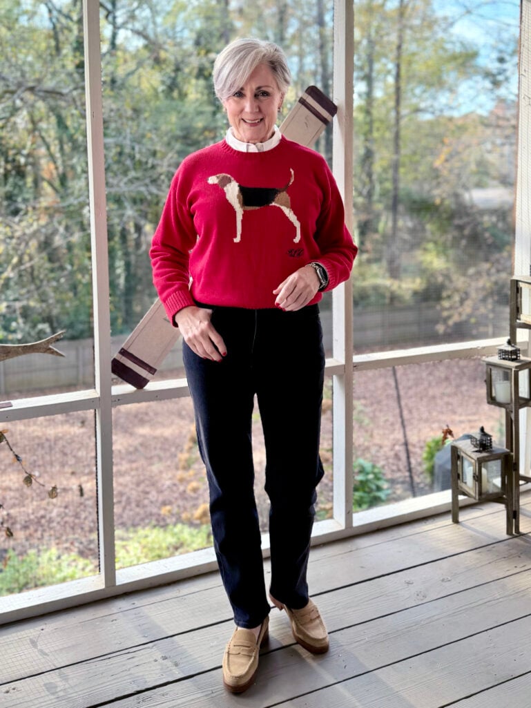 Beth in a red knit sweater with foxhound embroidery, white collar visible at neckline, dark jeans, and tan suede loafers, standing on a wooden porch