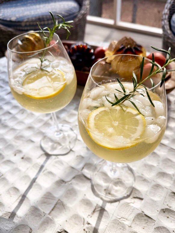 Two wine glasses filled with pale golden Rosemary Spritz cocktails garnished with lemon wheels and fresh rosemary sprigs, served over ice