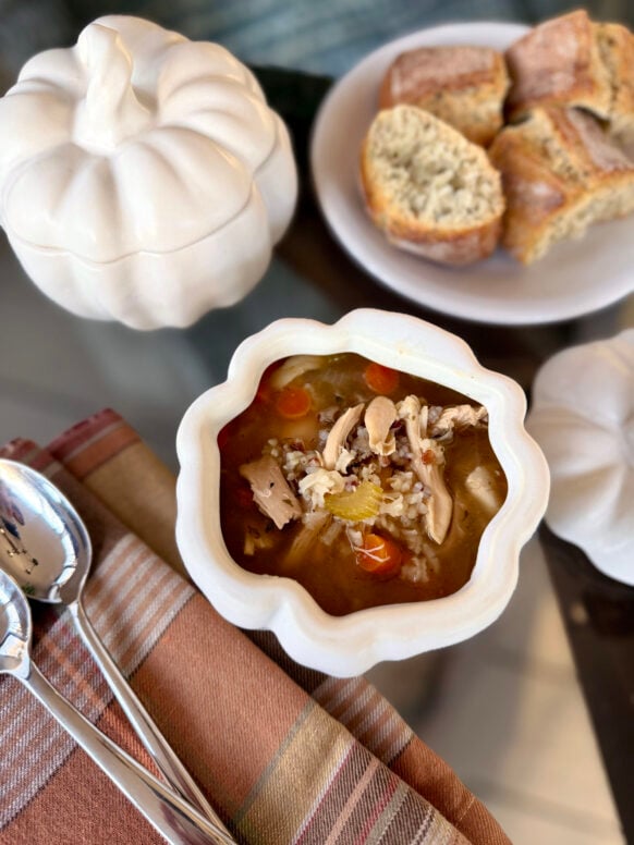 Overhead view of turkey wild rice soup in a white ceramic pumpkin-shaped bowl with shredded turkey, wild rice, carrots, and broth, served with crusty bread and plaid napkin