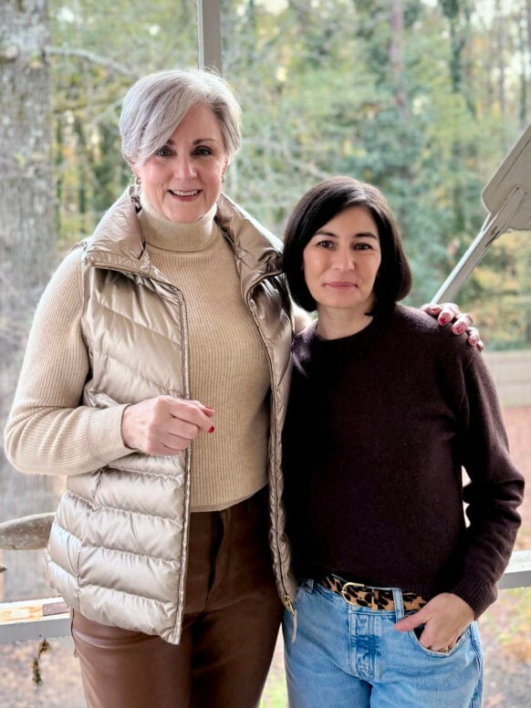 Two women standing together on a screened porch with natural fall lighting, one in a gold metallic vest over a camel turtleneck with faux leather pants, one in a brown cashmere sweater with barrel leg jeans and a leopard belt