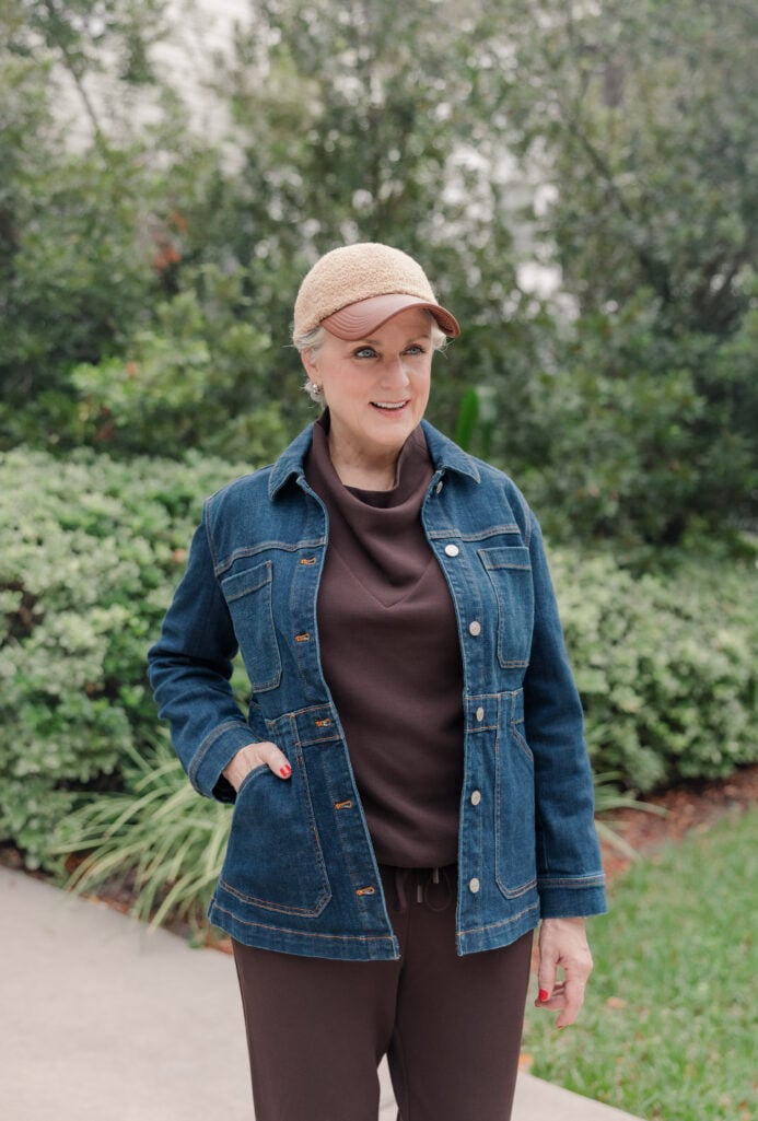 Beth wearing brown funnel-neck top and matching brown drawstring pants with dark wash denim jacket and tan sherpa baseball cap with leather brim, standing outdoors with lush green landscaping in background