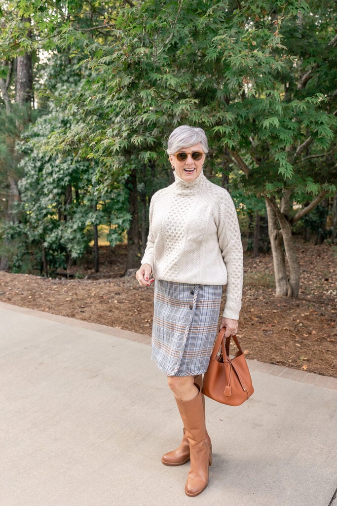 Beth Djalali wearing a cream cable-knit sweater with a plaid A-line midi skirt and cognac riding boots - preppy textured winter outfit