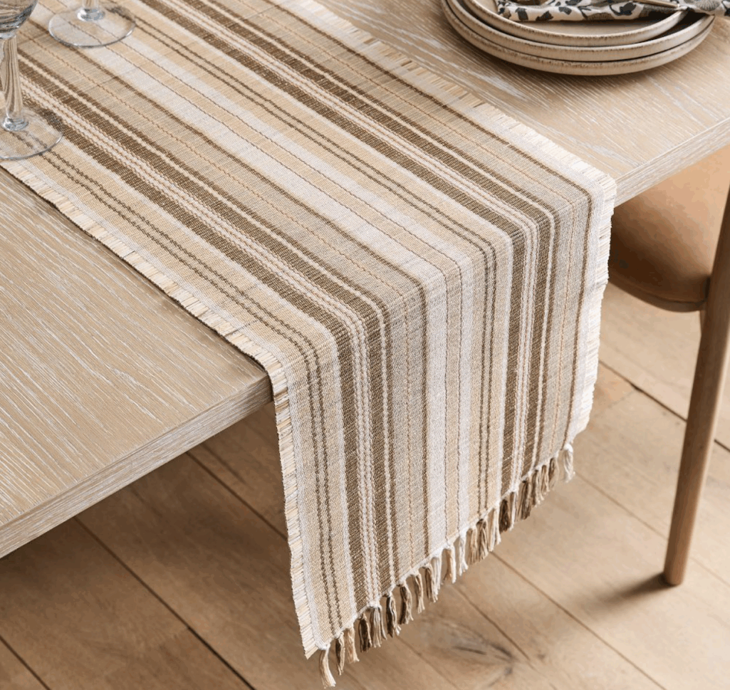 Natural fibers come together in this warm, neutral striped table runner with fringe details at the edges. Handwoven, 14-inches wide and 90-inches long. The different shades of tan and brown add warmth to any tablescape.