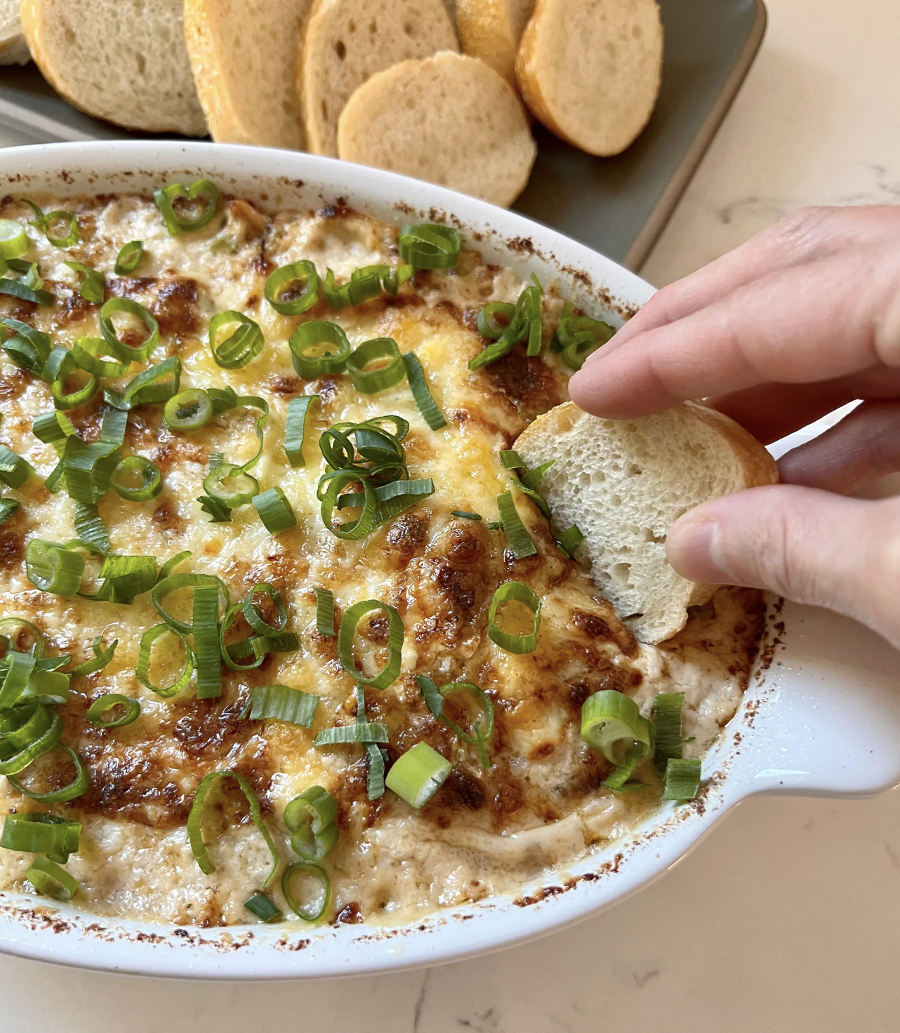 Kelly's hot and cheesy crab dip baked in a white dish, topped with fresh chopped spring onions and served with white bread crostini slices.