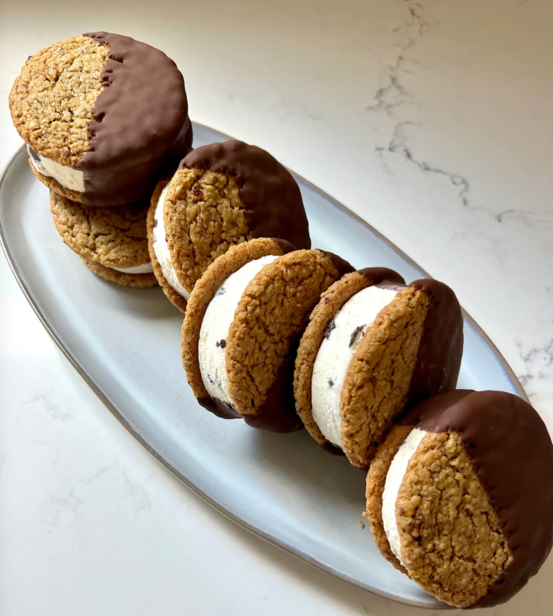 Plate of San Francisco–style ice cream sandwiches made with chewy oatmeal cookies and thick vanilla ice cream centers, each cookie half-dipped in chocolate and arranged in a neat row on a light gray platter.