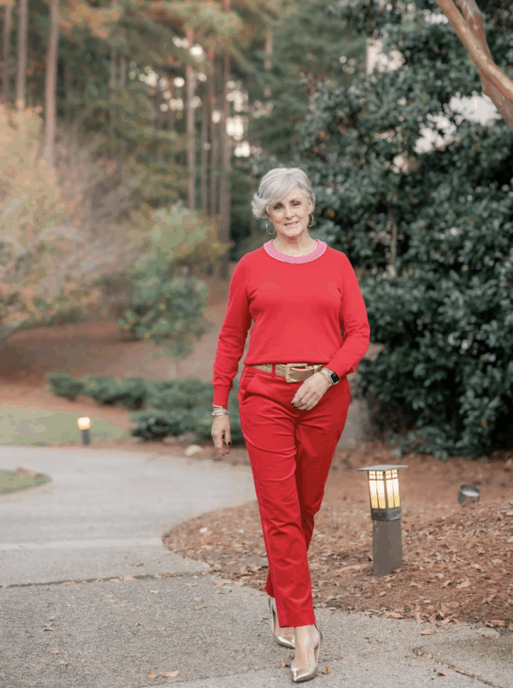 Woman with short silver hair wearing a red sweater and matching red pants, styled with a gold belt, hoop earrings, bracelets, and metallic heels, walking outdoors on a tree-lined path.