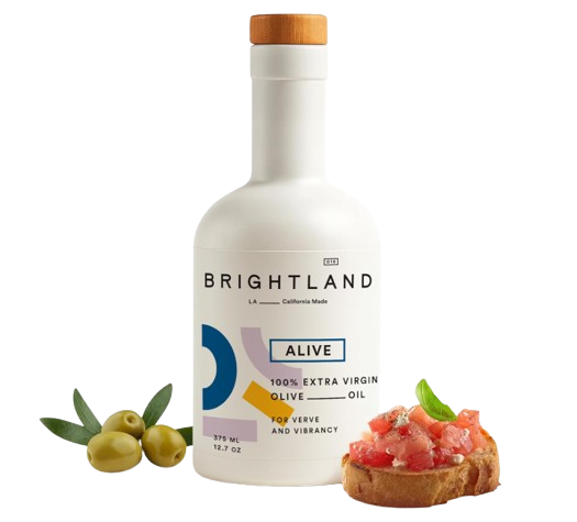 Brightland ‘Alive’ extra-virgin olive oil bottle with a white label and wooden cap, shown alongside fresh green olives and a tomato-topped crostini garnish.