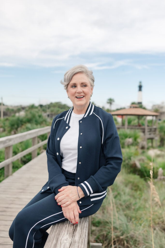 Beth wearing navy Spanx AirEssentials track suit with white piping detail and white crewneck underneath, seated on wooden boardwalk railing with lighthouse and coastal greenery in background